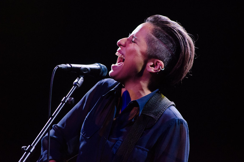 A young Latinx woman with short slicked-back hair is singing into a microphone with her eyes closed and er mouth open.