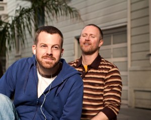 A man stands behind another man wearing headphones, trying to interrupt him, as he looks on into the distance