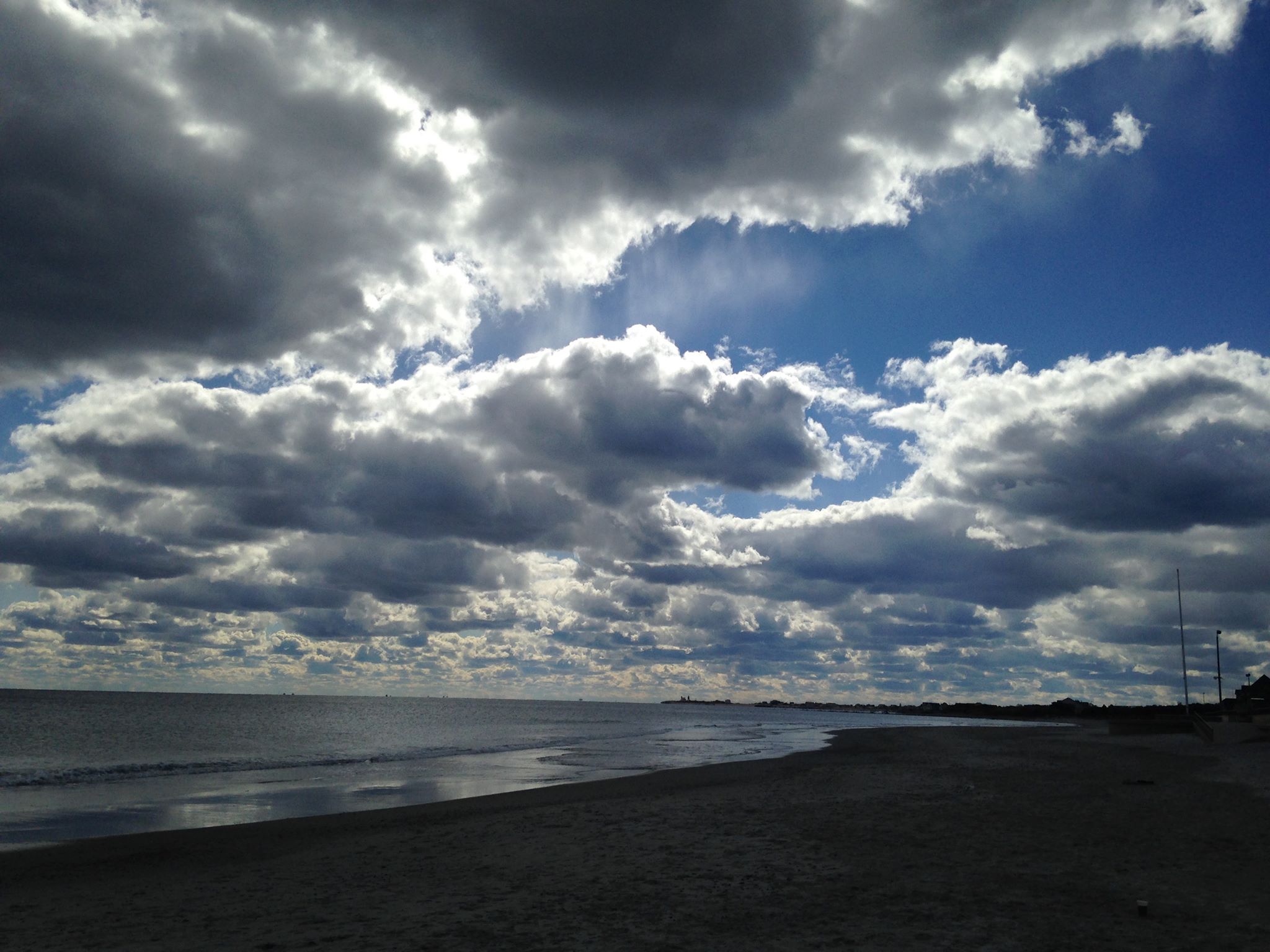 This is a photograph of a view of the Scarborough State Beach at Newport, Rhode Island. The sky is a dark, bright blue with sun illuminating big, bright, puffy clouds. The tide is out, the water is calm, and the sand is darkened.