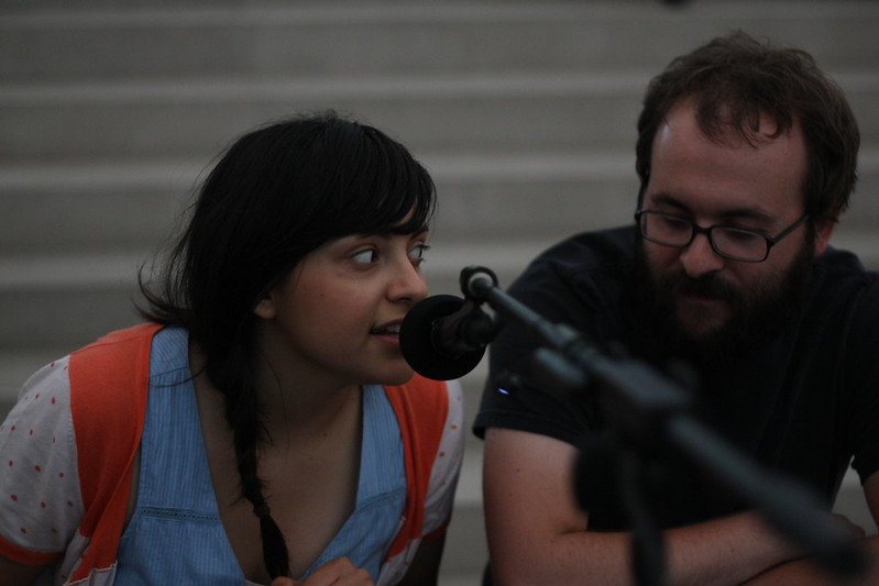 A young woman in an orange and blue outfit with long dark hair worn in one braid over her shoulder speaks into a microphone, while a young man with a beard and glasses looks sits next to her. quiet and looking down.