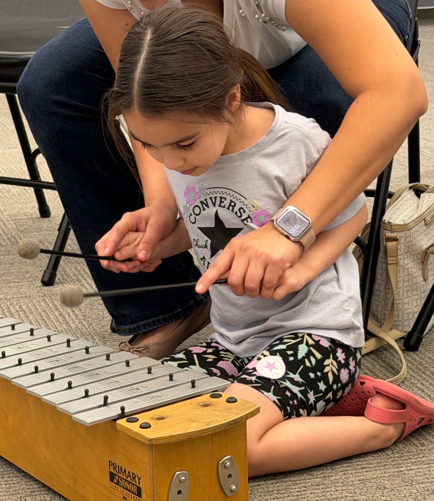 A young Latine girl dressed in brightly colored patterned shorts and a matching T-shirt, sits on her knees over a xylophone. She holds the mallets, along with a woman who sits over her, helping her find the notes. Only the woman's torso and legs are visible